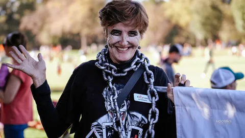 A woman in a chain necklace costume smiles beside a banner in the park.