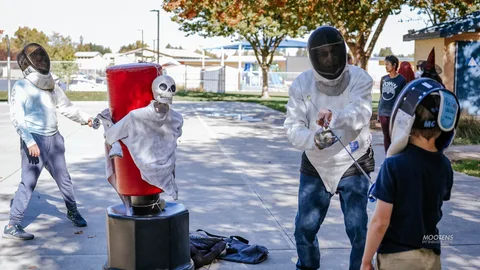 Two people in fencing masks spar around a skeleton-topped practice dummy.