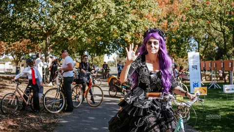 A cyclist in purple hair and sugar-skull makeup waves and smiles beside her bike in the fall foliage.