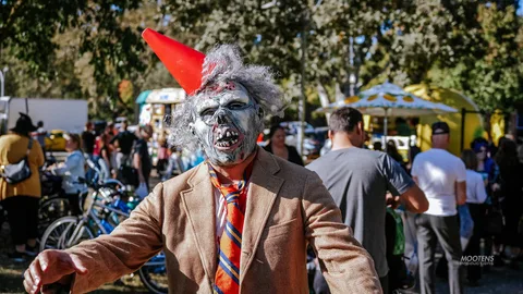 A man in an elderly zombie mask and red party hat strides through the festival food vendors.