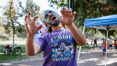 A man in zombie face paint snarls with claw hands raised, wearing a Zombie Bike Ride shirt.