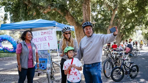 A family takes a selfie together beside a hand-painted zombie voting sign, bikes parked nearby.