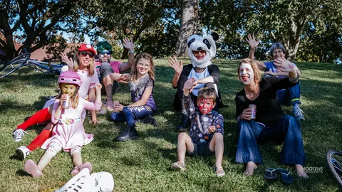 A child with face paint sits on the grass with family nearby.