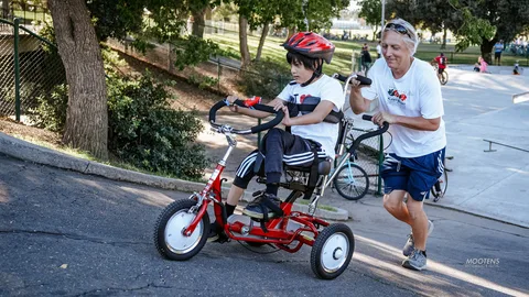 A boy on a red adaptive trike is guided along the path by a volunteer running beside him.