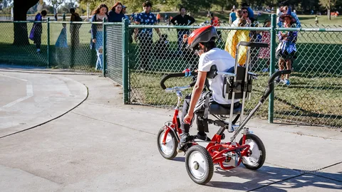 A costumed rider sits on a red adaptive trike near the fence line.