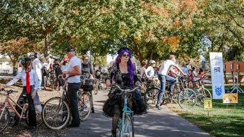 A cyclist in purple-haired sugar-skull makeup and lace rides toward the camera through a busy costumed crowd.