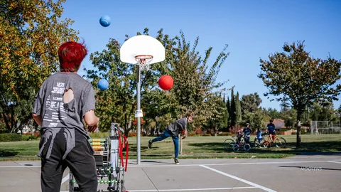 A teen shoots a basketball on an outdoor court near a small wheeled robot.