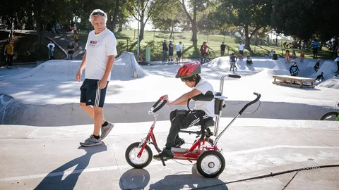 A boy on a red adaptive trike waits at the edge of the skate park.