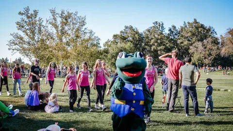 The frog mascot salutes alongside a family in the park.
