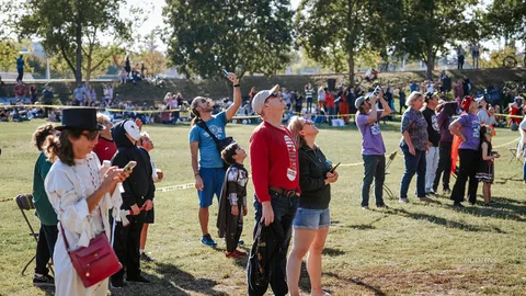 A long line of costumed spectators looks up together, watching the skydivers descend.