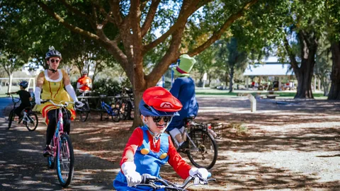 A child in a Mario costume rides a bike through the park with a Princess Peach costumed rider behind.
