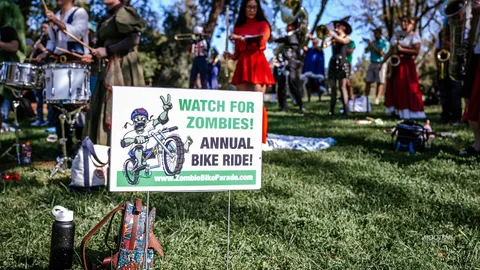 A Watch for Zombies annual bike ride sign stands in the grass in front of a costumed marching band.