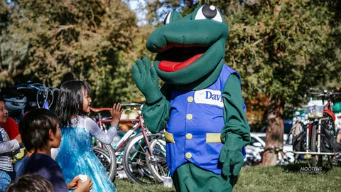 A green frog mascot in a Davis vest waves and high-fives delighted kids.