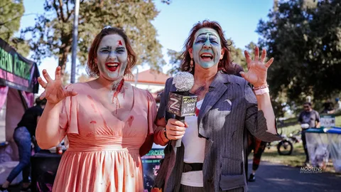 Two women in ghoulish zombie makeup laugh and pose with a Zombie News Now prop microphone.
