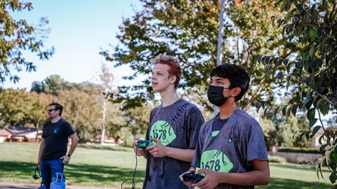 Two teens hold robot controllers wearing matching competition jerseys.