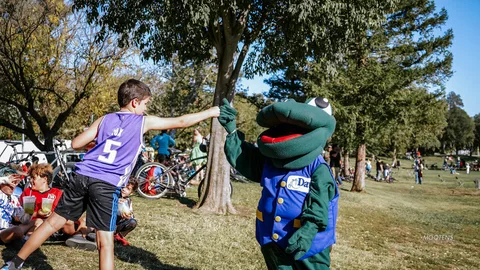 A boy fist-bumps the green frog mascot under a shady tree.