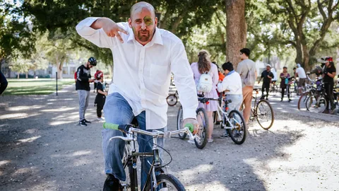 A man in ghoulish face paint raises his hands while walking his bike down a tree-lined path.