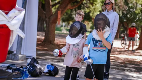 Two kids in fencing masks and gear pose together with a parent.