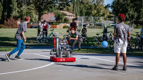 Teens play basketball on an outdoor court beside a small wheeled robot.