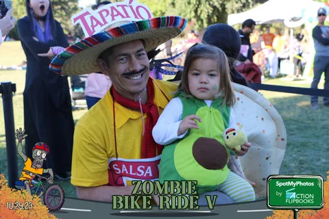 A man in a sombrero costume holds a smiling child dressed as an avocado.