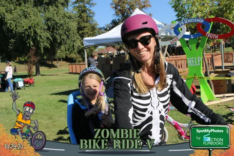 A girl in a pink helmet stands near a booth with a woman in skeleton costume.