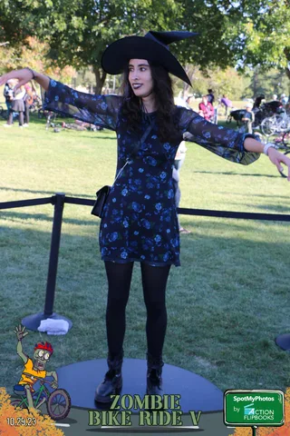 A woman in a witch hat and dress poses dramatically with arms spread wide, closer crop.