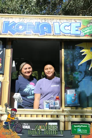 Two women smile from inside a festival ticket booth window.