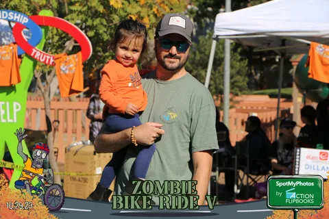 A man holds a toddler near a colorful festival booth sign.