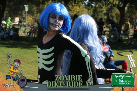 A woman in a skeleton costume and blue wig raises her arm triumphantly.