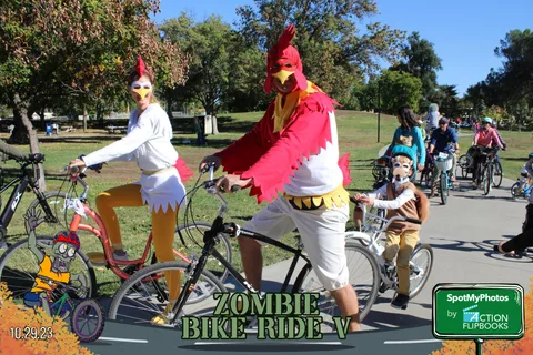 Two riders in full chicken costumes lead a line of cyclists along the park path.