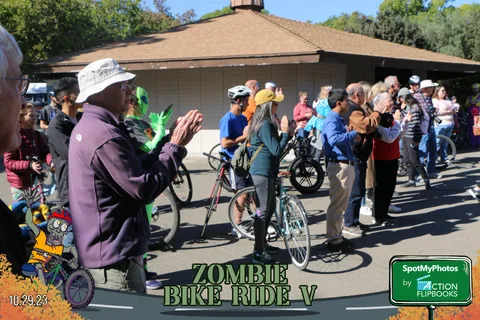 A performer in face paint and a purple jacket stands as cyclists gather nearby.