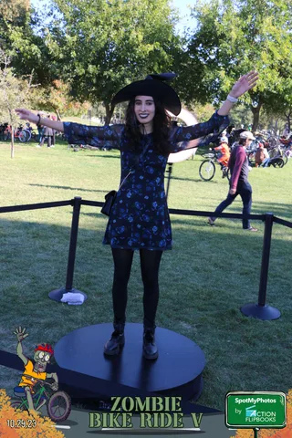 A woman in a witch hat and dress strikes a dramatic pose with arms outstretched.