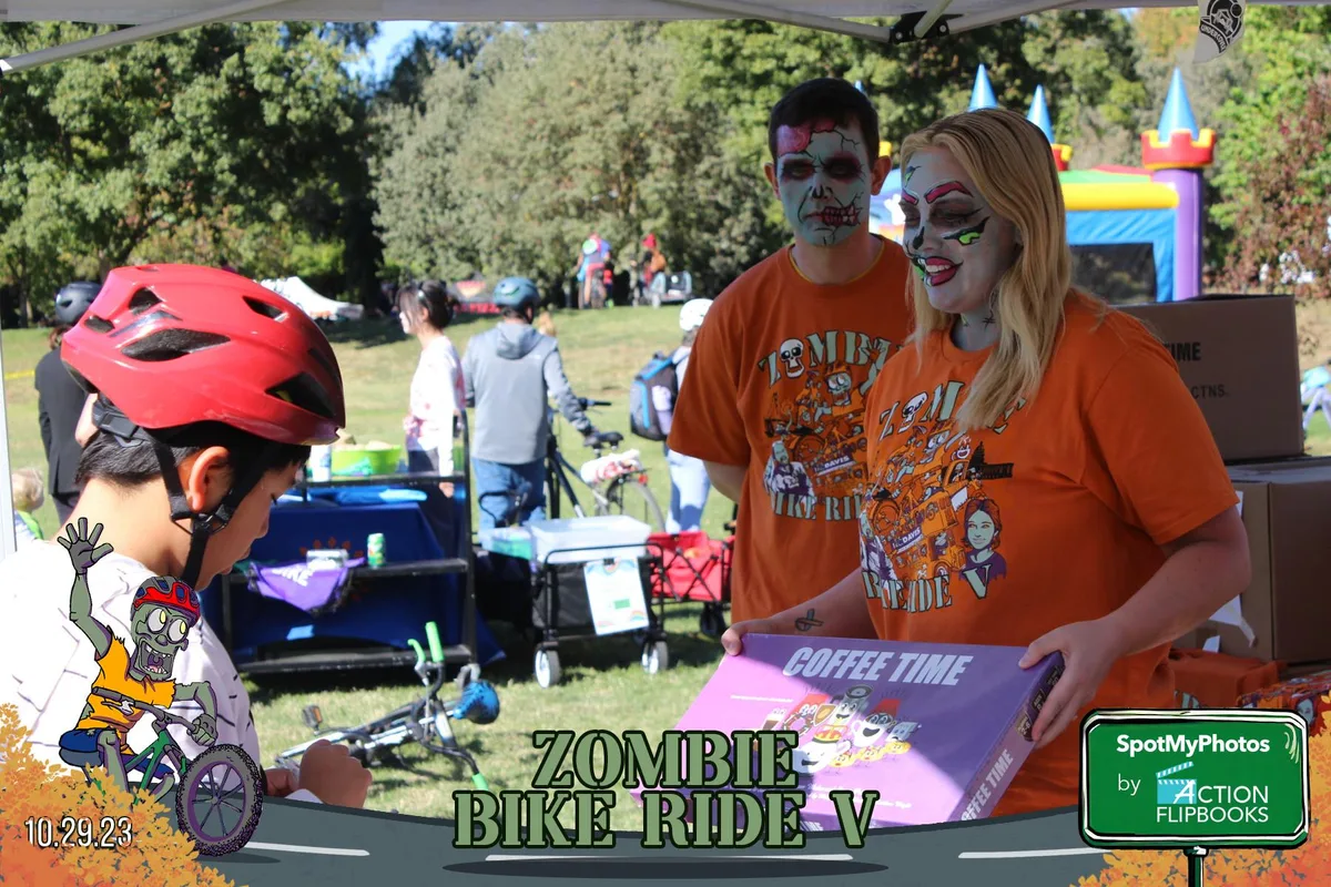Zombie-painted volunteers in orange shirts hand a Coffee Time board game to a helmeted boy.