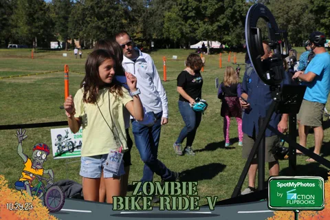 A girl waves near photo booth equipment while adults talk in the background.