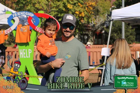 A man holds a young child in an orange shirt near festival signage.