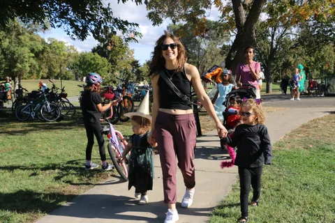 A smiling woman walks hand in hand with two costumed kids along a park path.