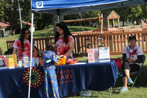 Blood-splattered volunteers staff the Yolo County Library booth beside a Davis Makerspace sign.