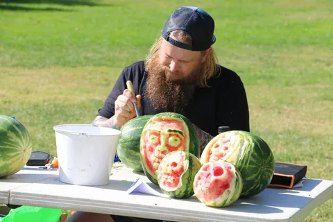 A bearded artist carves skull faces into watermelons at his festival booth.