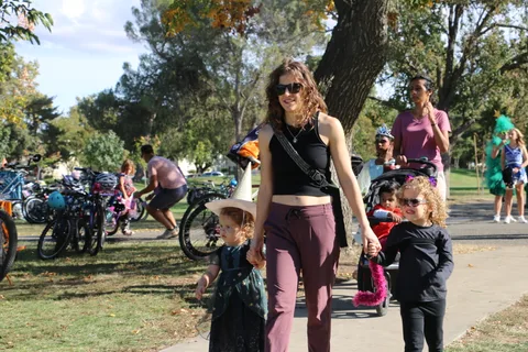 A woman walks hand in hand with two costumed toddlers along a tree-lined path.