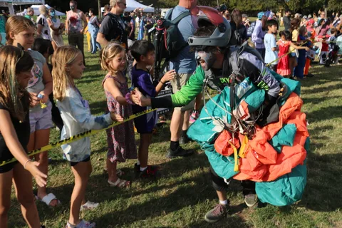 Kids reach through a tape line to greet a costumed skydiver holding a folded parachute.