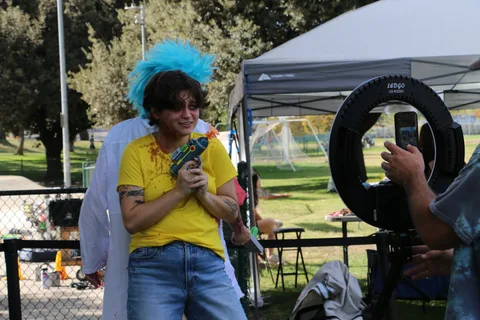A man in a blue mohawk wig strikes a playful pose with a neon toy gun.