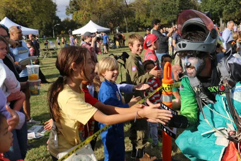 A costumed skydiver in zombie face paint high-fives a line of laughing kids at the finish tape.