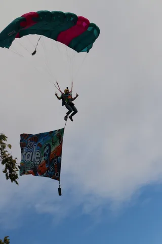 A tandem skydiver descends with an illustrated banner unfurled beneath the canopy.