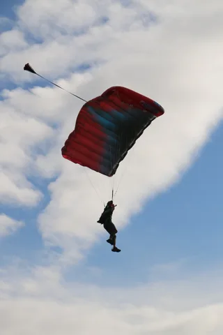 A skydiver under a red canopy glides across a blue sky with scattered clouds.