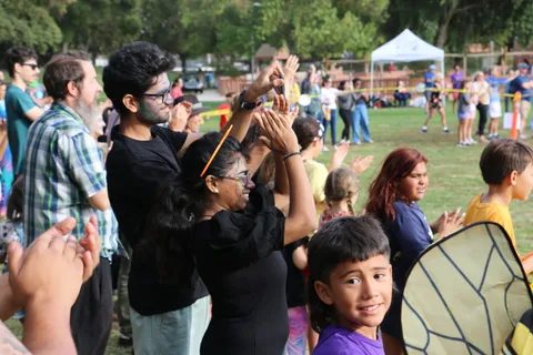 Kids in costume, including one in butterfly wings, clap and cheer watching the skydivers land.