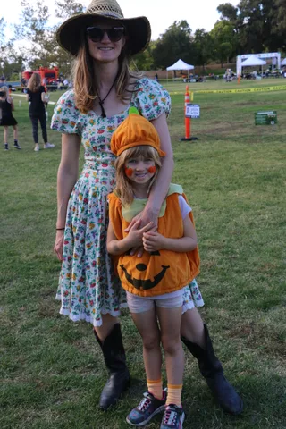A mother in a floral dress holds her daughter, dressed as a pumpkin, both smiling widely.