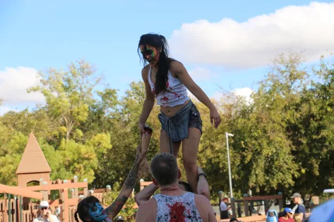 A zombie-painted performer leaps mid-air across the grass near the playground.