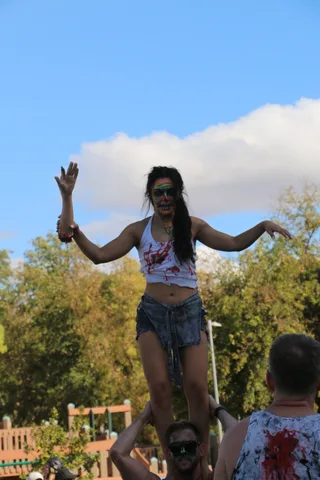 A zombie-painted performer claws toward the camera mid-dance against a clean blue sky.