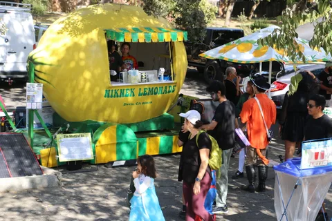A costumed child stands near the bright yellow Mikuni Lemonade stand.
