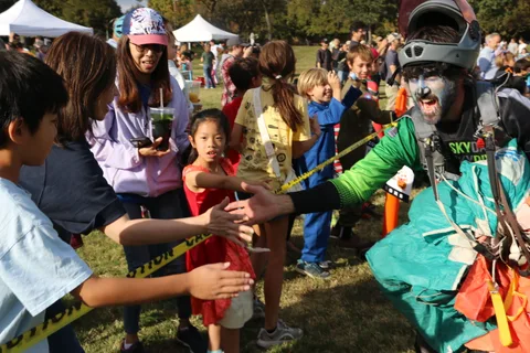 Kids in costume reach up to greet a skydiver in zombie face paint behind a caution tape line.